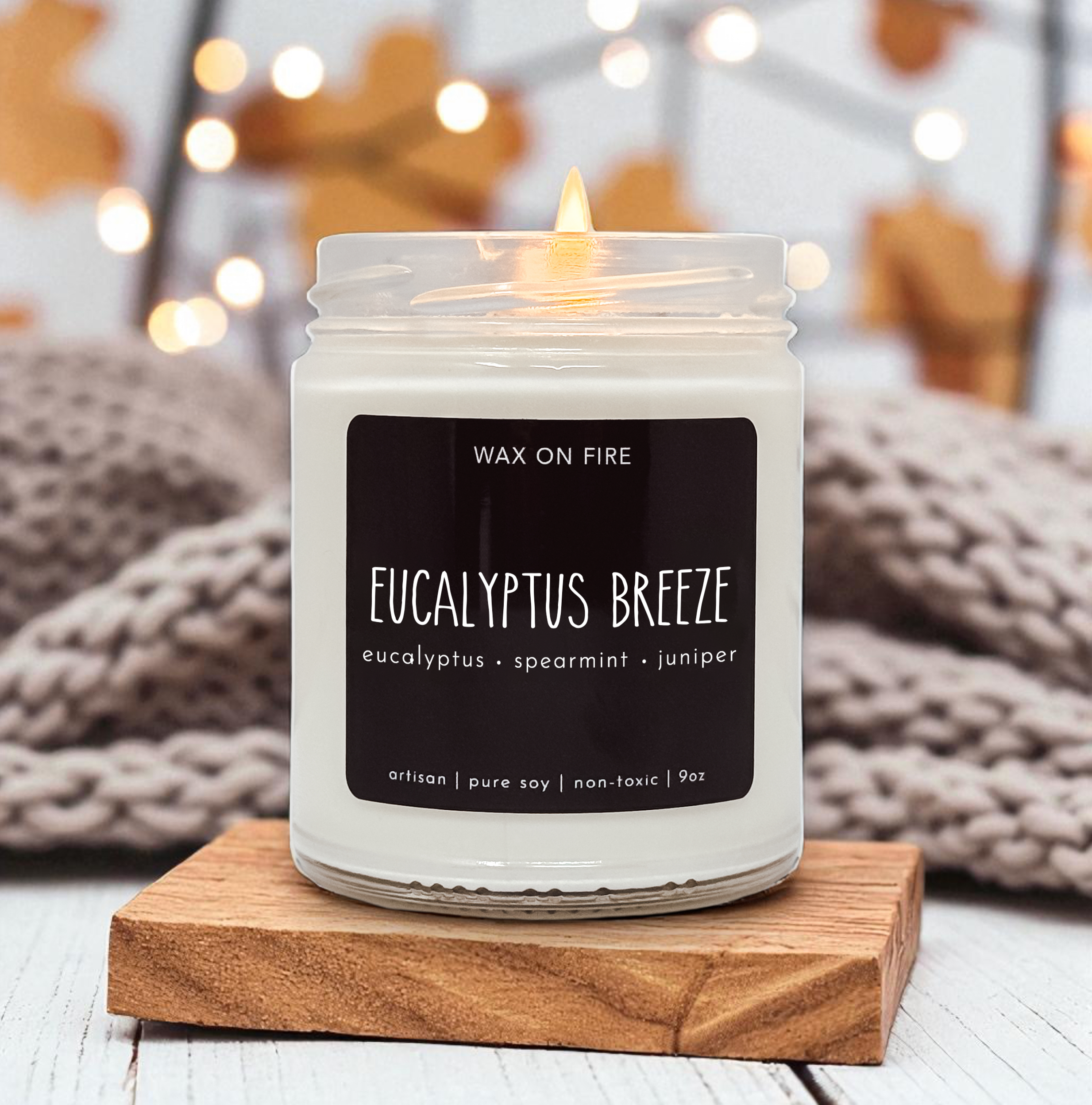 A white candle labeled "Eucalyptus Breeze" sits on a wooden coaster, with a blurred background featuring a cozy, warm atmosphere.