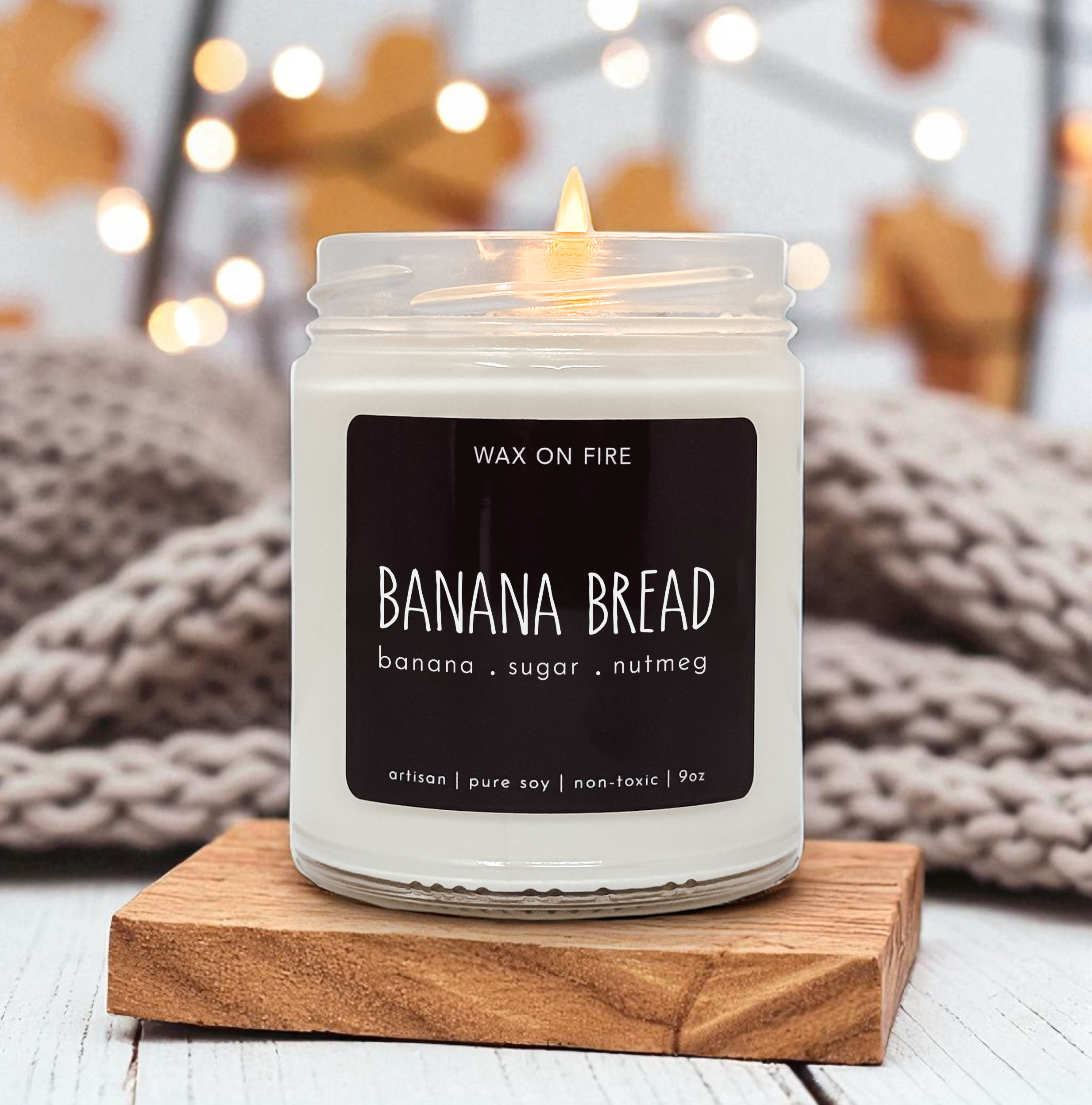 A white candle labeled "Banana Bread" sits on a wooden coaster, with a blurred background featuring a cozy, warm atmosphere.