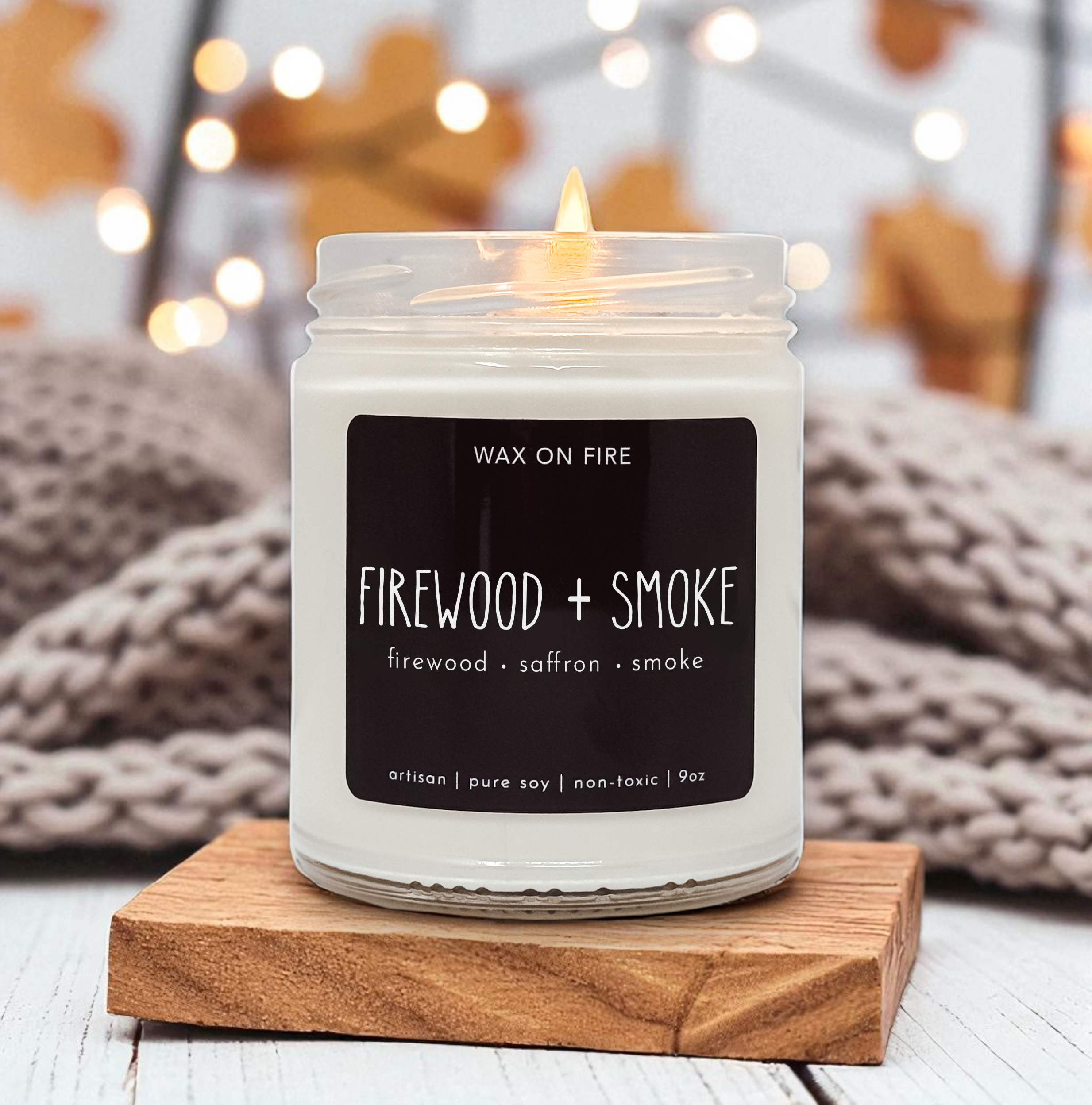 A white candle labeled "Firewood + Smoke" sits on a wooden coaster, with a blurred background featuring a cozy, warm atmosphere.
