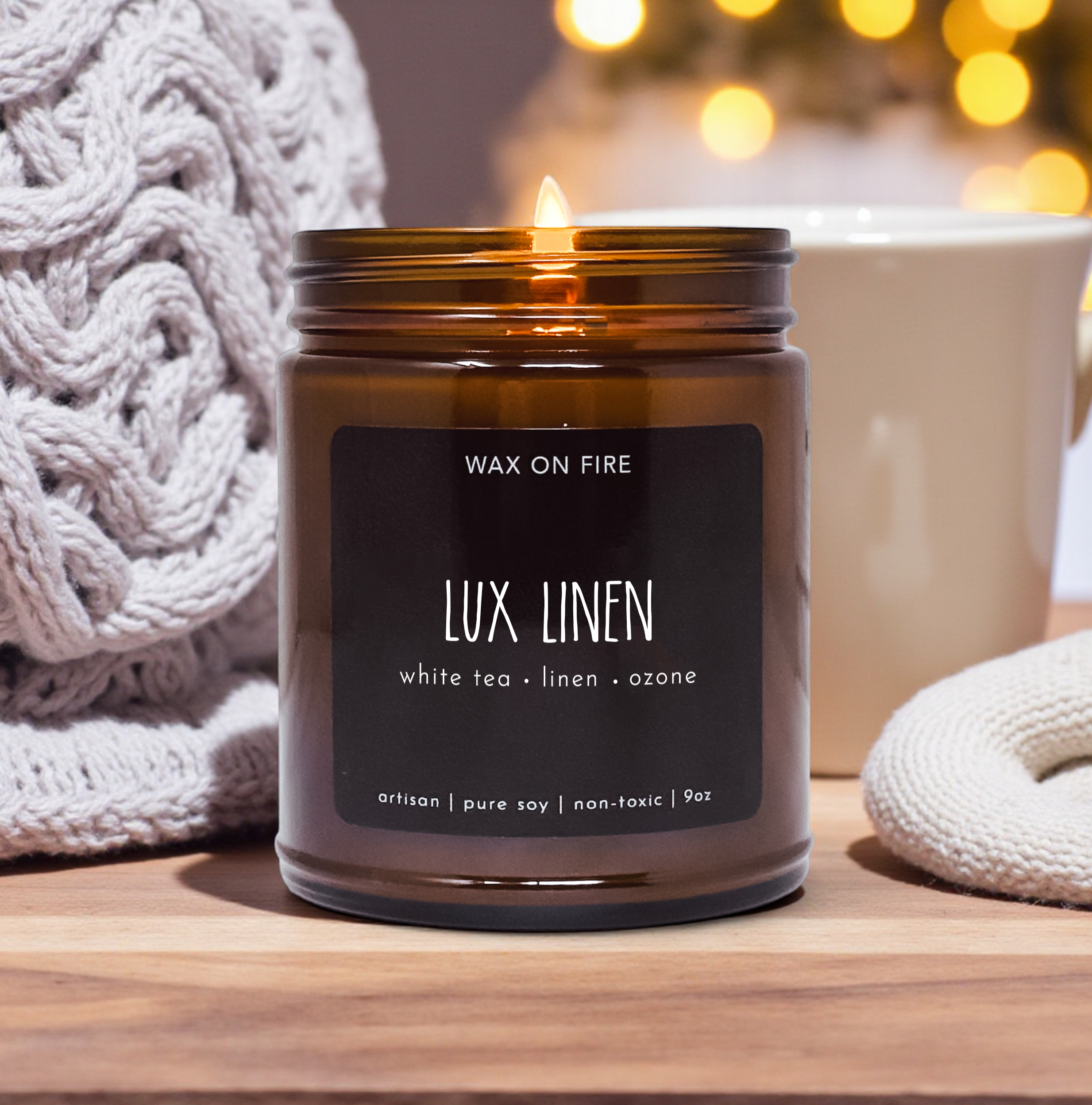 A brown glass jar candle labeled "Wax on Fire LUX LINEN" sits on a wooden surface, with a white mug and a white knitted item in the background.