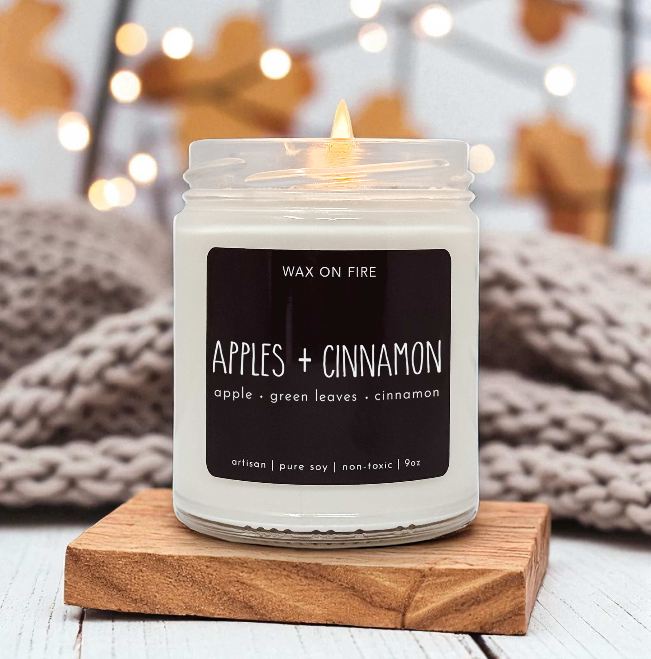 A white candle labeled "Wax on Fire" sits on a wooden coaster, with a blurred background featuring a cozy, autumnal setting.